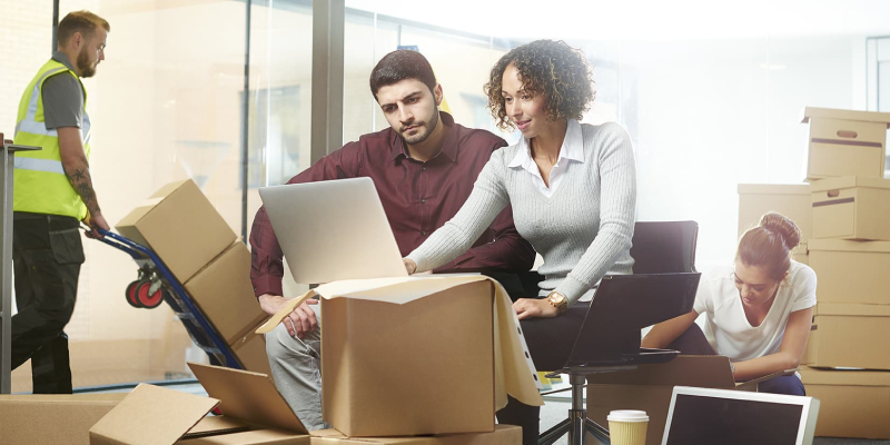 two employees are working on the laptop while there are other two persons packing office things for relocating them.